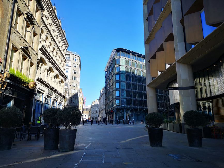 A city street scene with a mix of historic and modern buildings. On the left, an ornate, classical-style facade with detailed stonework and decorative elements, partially shaded by large potted shrubs