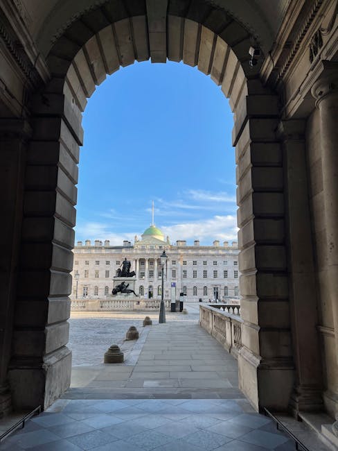 View through a stately stone archway onto the courtyard of Somerset House, featuring neatly maintained garden beds and a paved pathway leading towards a large, historic building with a green domed roo