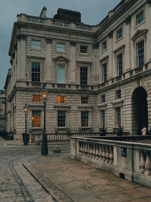View through a stately stone archway onto the courtyard of Somerset House, featuring neatly maintained garden beds and a paved pathway leading towards a large, historic building with a green domed roo
